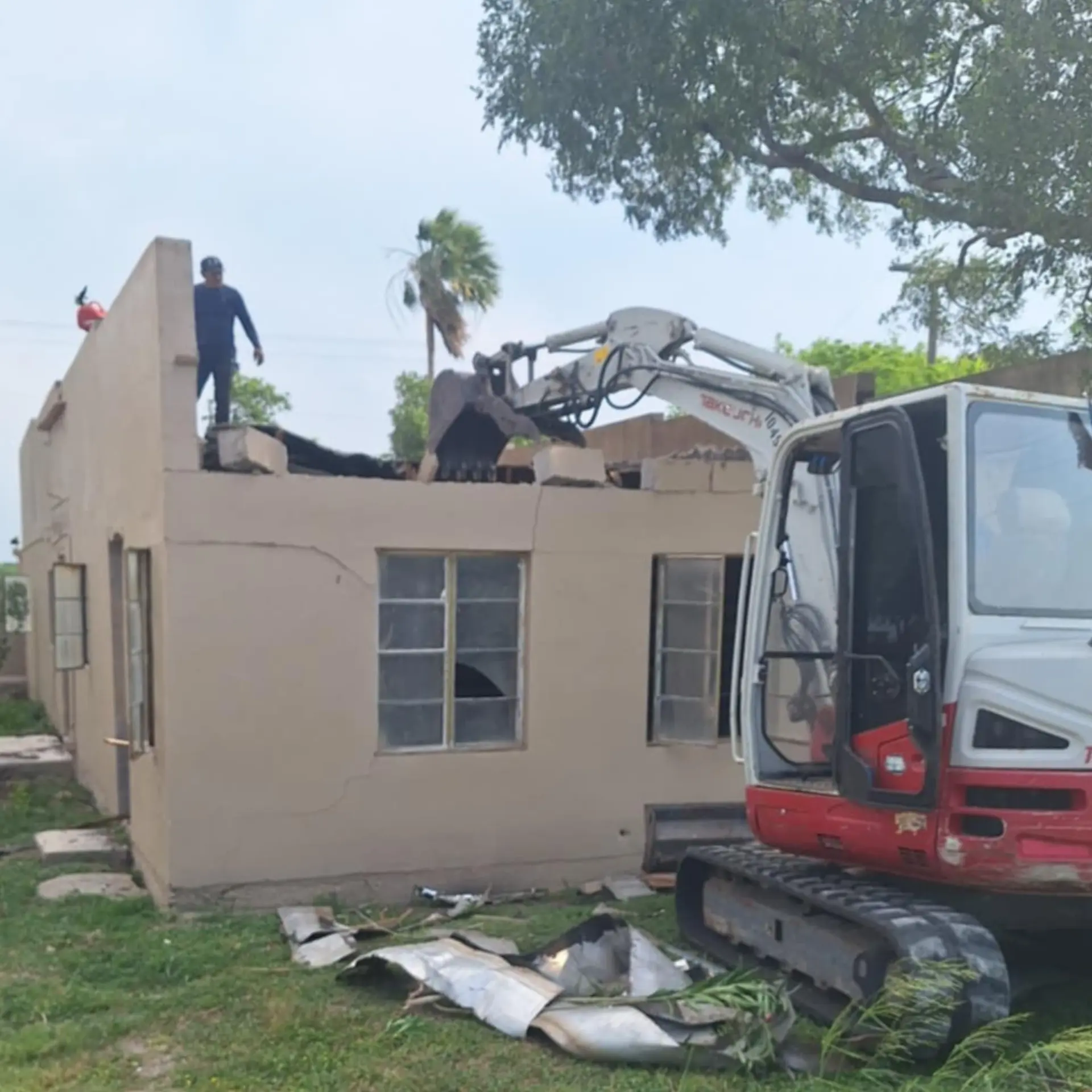 Excavator removing the roof of a masonry house while a worker stands on the wall